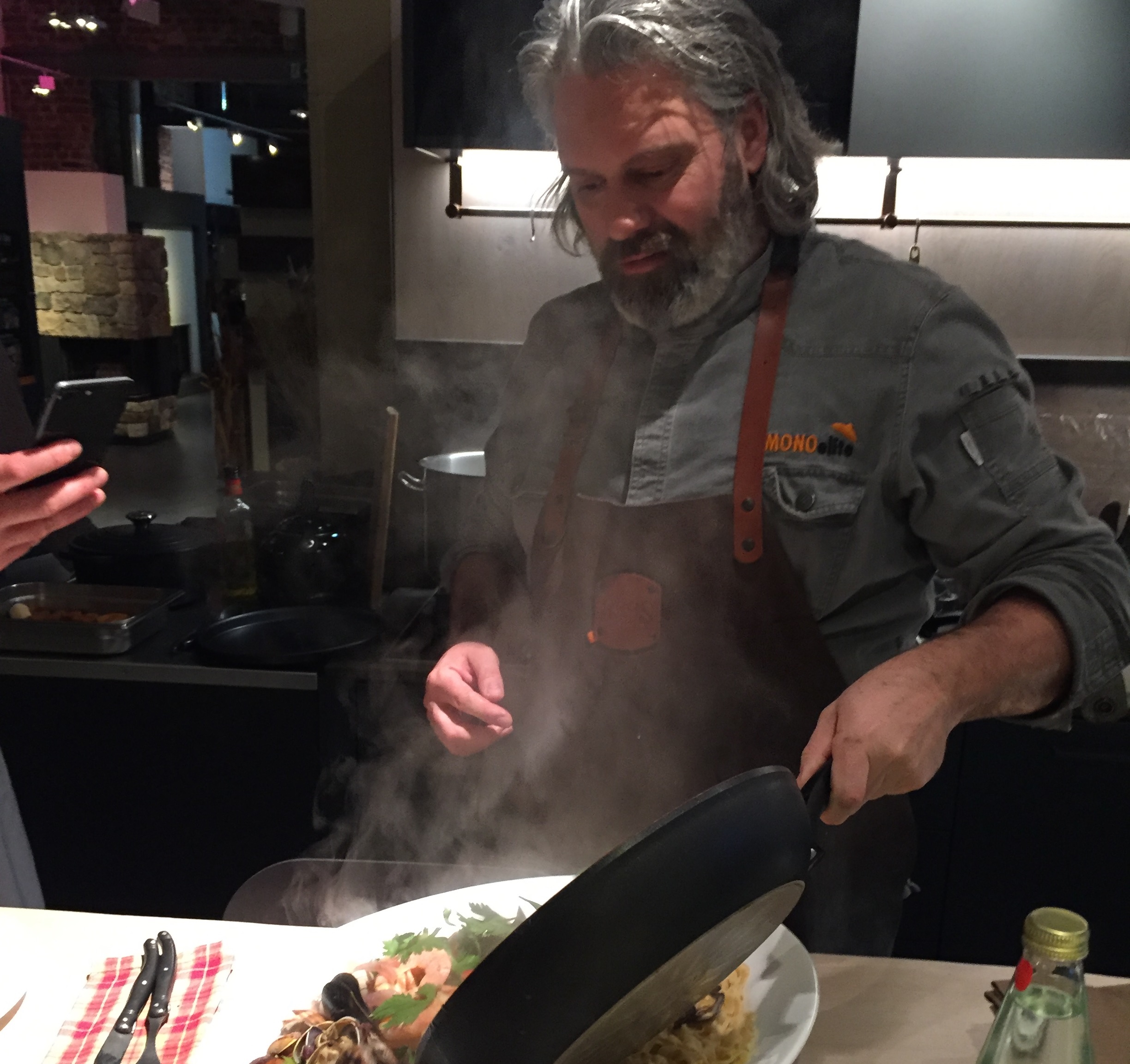 Koch mit grauem Bart serviert dampfendes Gericht aus Pfanne, trägt Schürze und steht in moderner Küche mit dunklem Design.