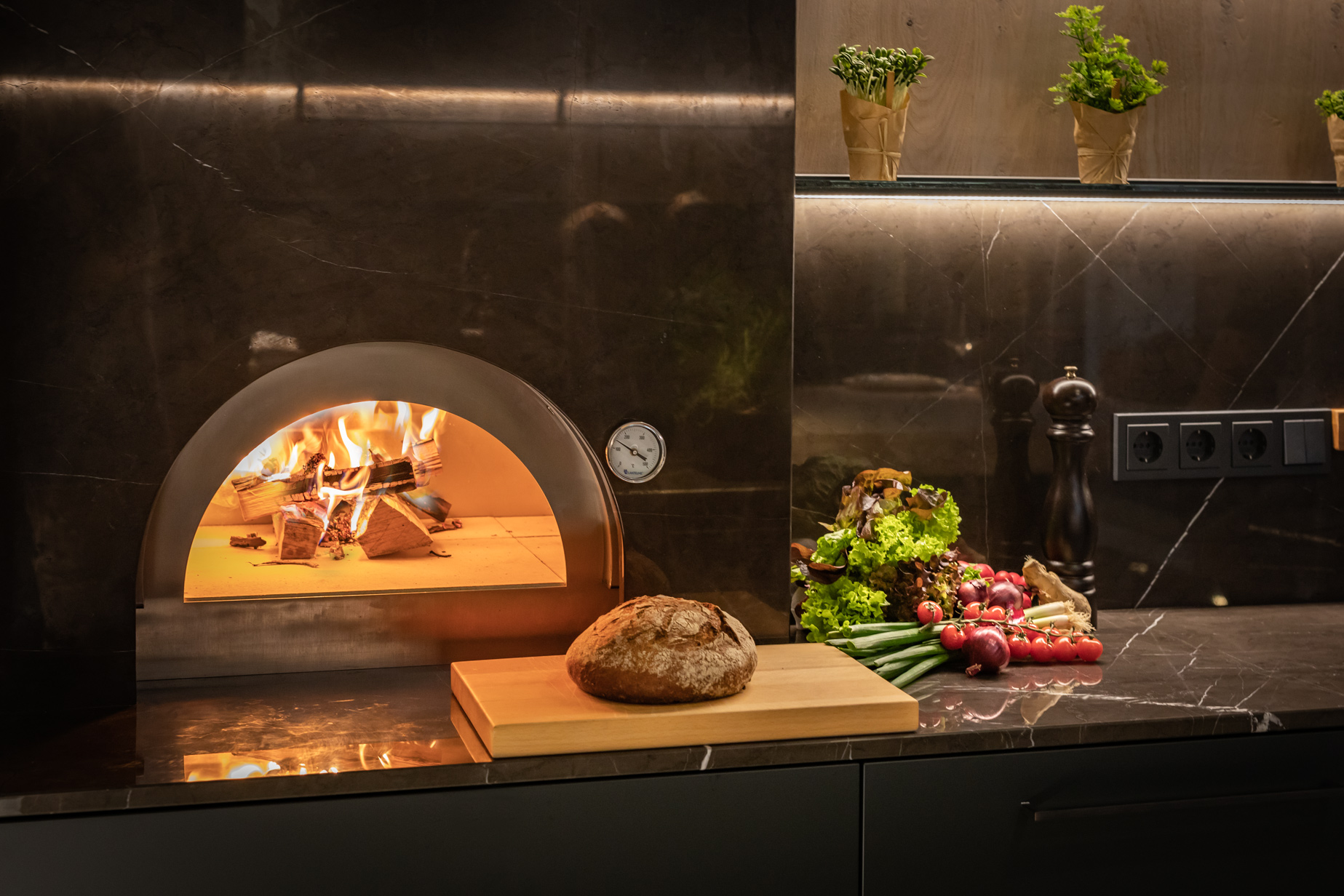 Holzofen mit Flammen, frisches Brot auf Schneidebrett, daneben Gemüse und Kräuter auf dunkler Küchenarbeitsfläche.