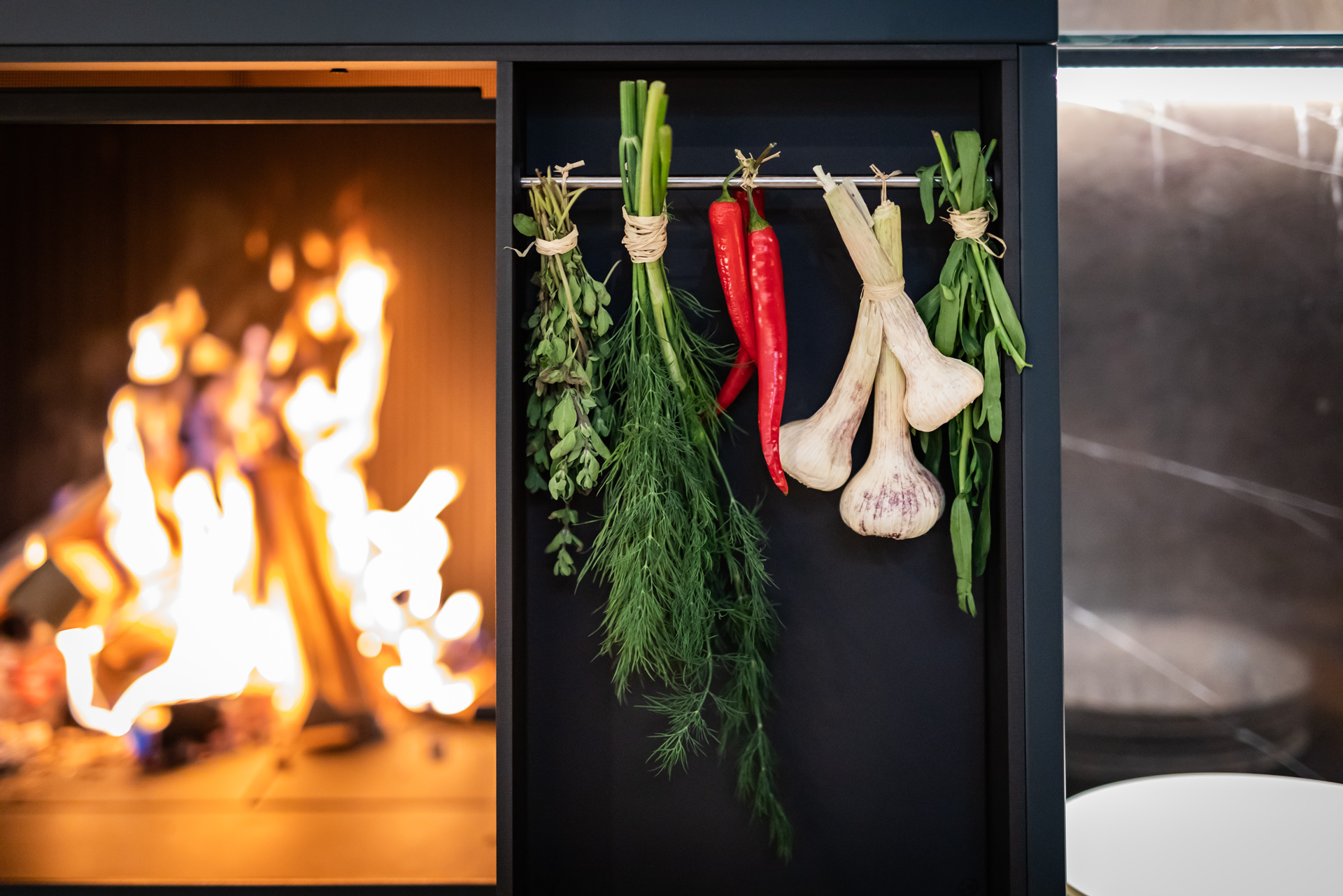 Kräuter, Chili und Knoblauch hängen dekorativ neben einem offenen Feuer in einer modernen Küche mit stilvollem Design.