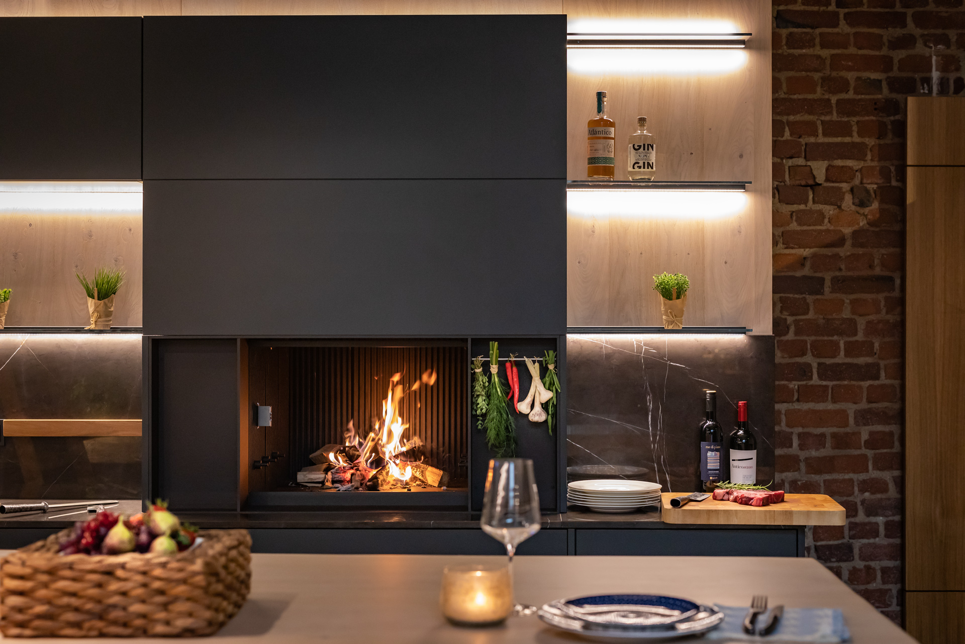 Moderne Küche mit integriertem Kaminfeuer, stimmungsvoller Beleuchtung, Weinflaschen und frischen Zutaten auf der Arbeitsplatte.