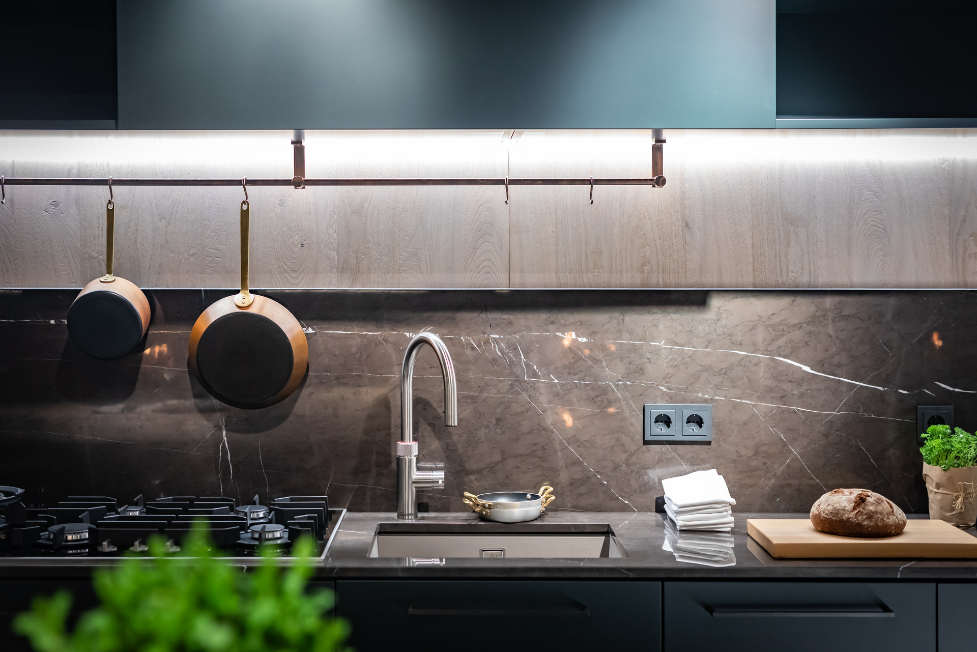 Moderne Küchenzeile mit Spüle, Gasherd, Kupfertöpfen, Brot und frischen Kräutern auf edler Stein-Arbeitsplatte.