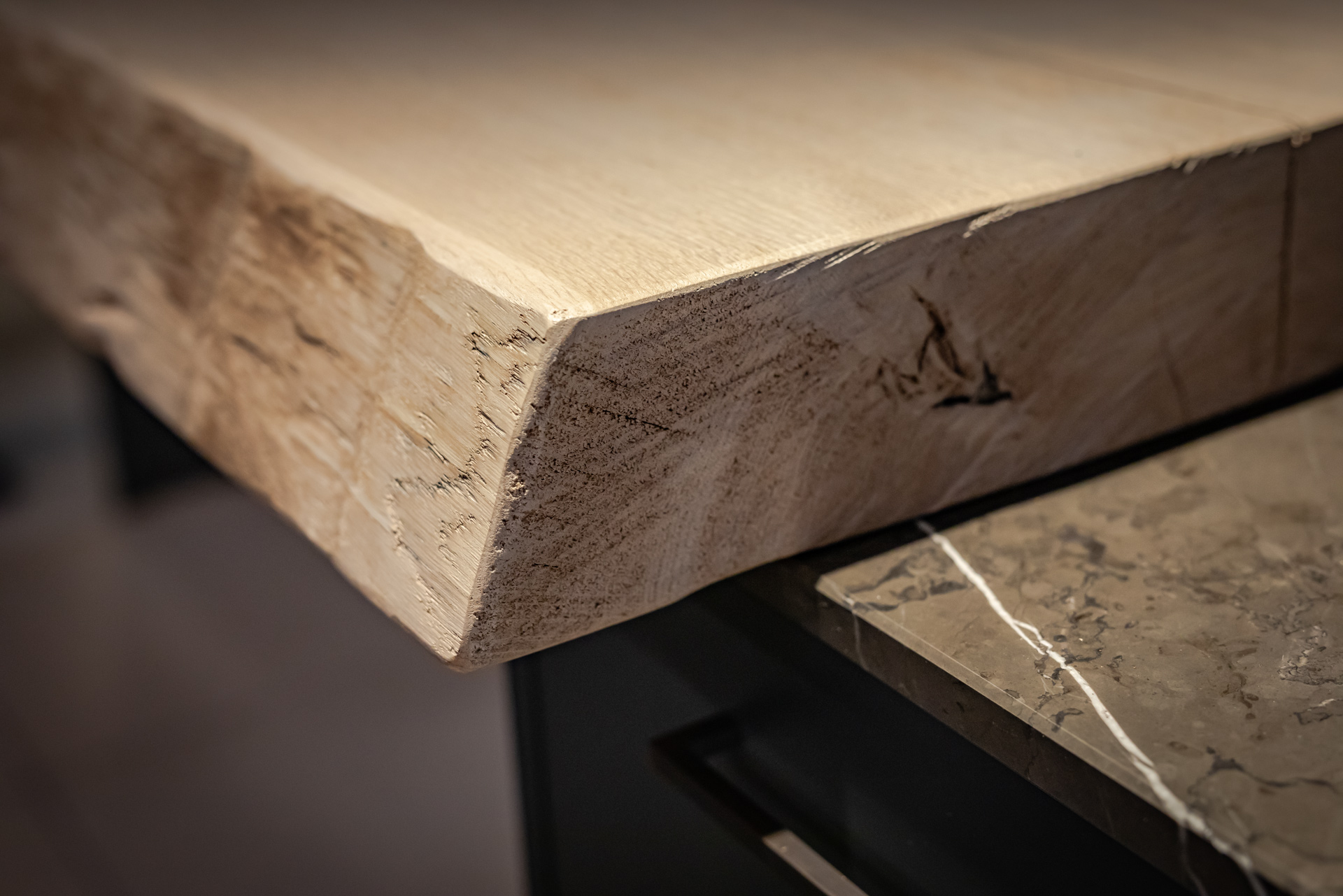 Detailaufnahme einer massiven, naturbelassenen Holztischkante über einer eleganten Küchenarbeitsplatte aus Stein.