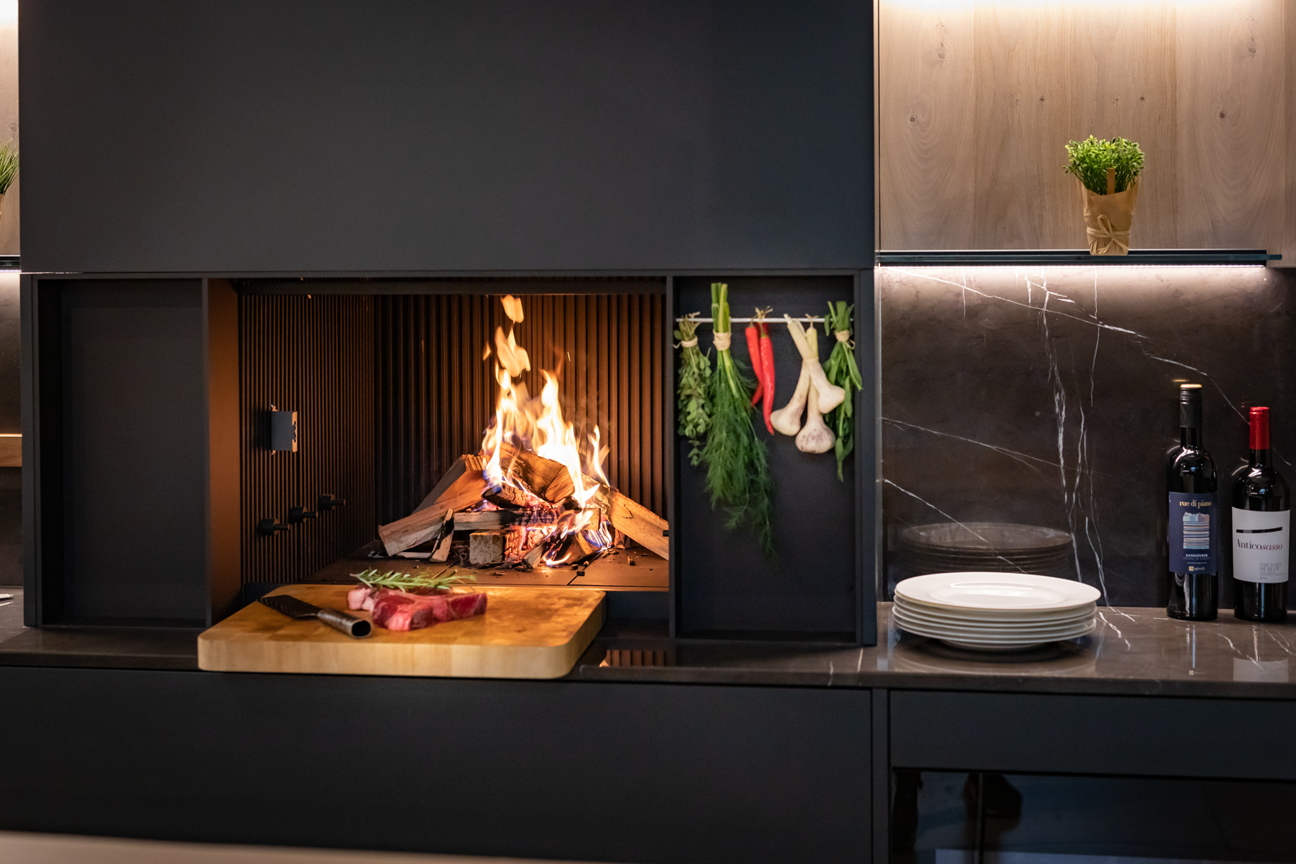 Offener Küchenherd mit Feuer, frischen Kräutern, Knoblauch, Fleisch auf Holzbrett und Weinflaschen im eleganten Ambiente.