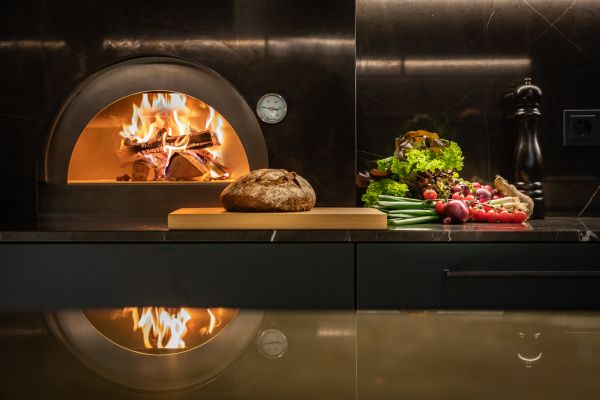 Feuer im Pizzaofen mit Brot auf Holzbrett und frischem Gemüse auf schwarzer Küchenplatte vor dunklem Marmorhintergrund.