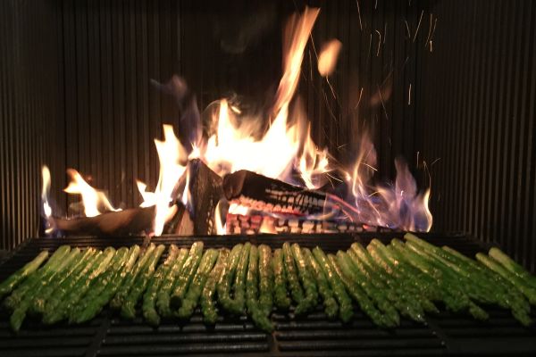 Grüner Spargel wird auf einem Grill über offener Holzflamme schonend gegart – Funken fliegen im Hintergrund.