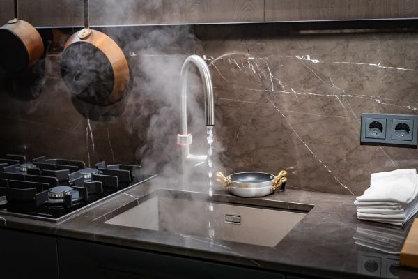 Moderne Küche mit Wasserhahn, aus dem Dampf aufsteigt, daneben schwarze Kochfelder und kupferfarbene Hängeschalen.