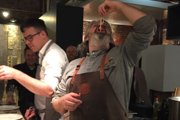 Koch kostet Pasta direkt aus dem Topf, während Gäste in geselliger Küchenatmosphäre zusehen und mitlachen.